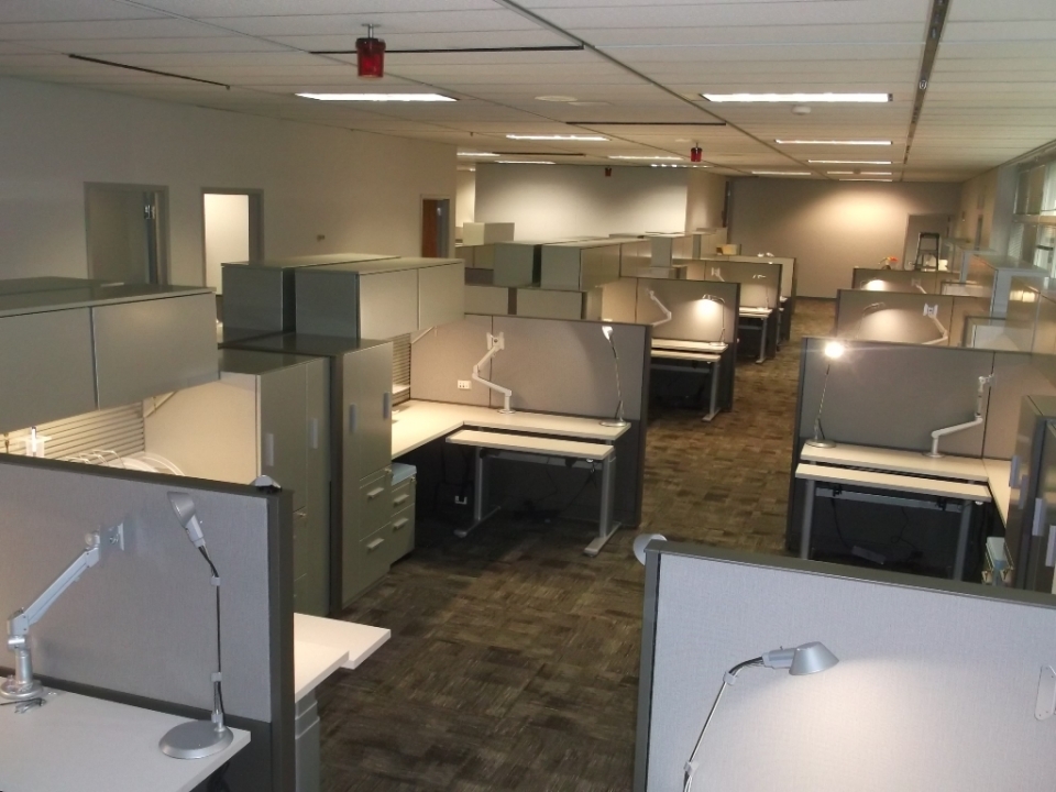 Photo of interior of office with cubicles. Each cubicle featuring lights, computer monitor mounts, and file cabinets