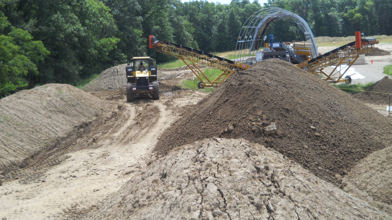 Photo at Iowa Army Ammunition Plant of environmental remediation work with piles of dirt, construction equipment, and the frame of an arched building in the background