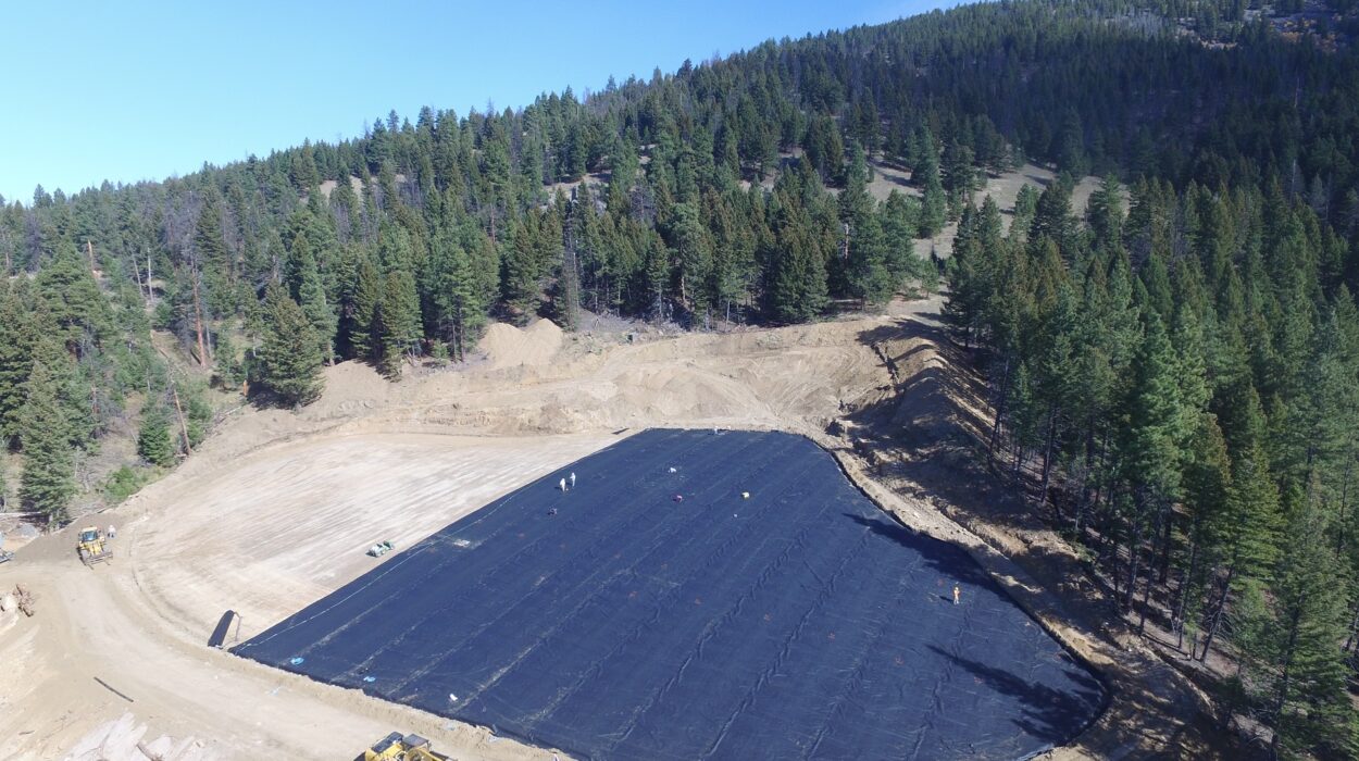 Aerial of a reclamation site in Montana with a lot of evergreen trees in the background and a large dirt area in the foreground partially covered in black covering