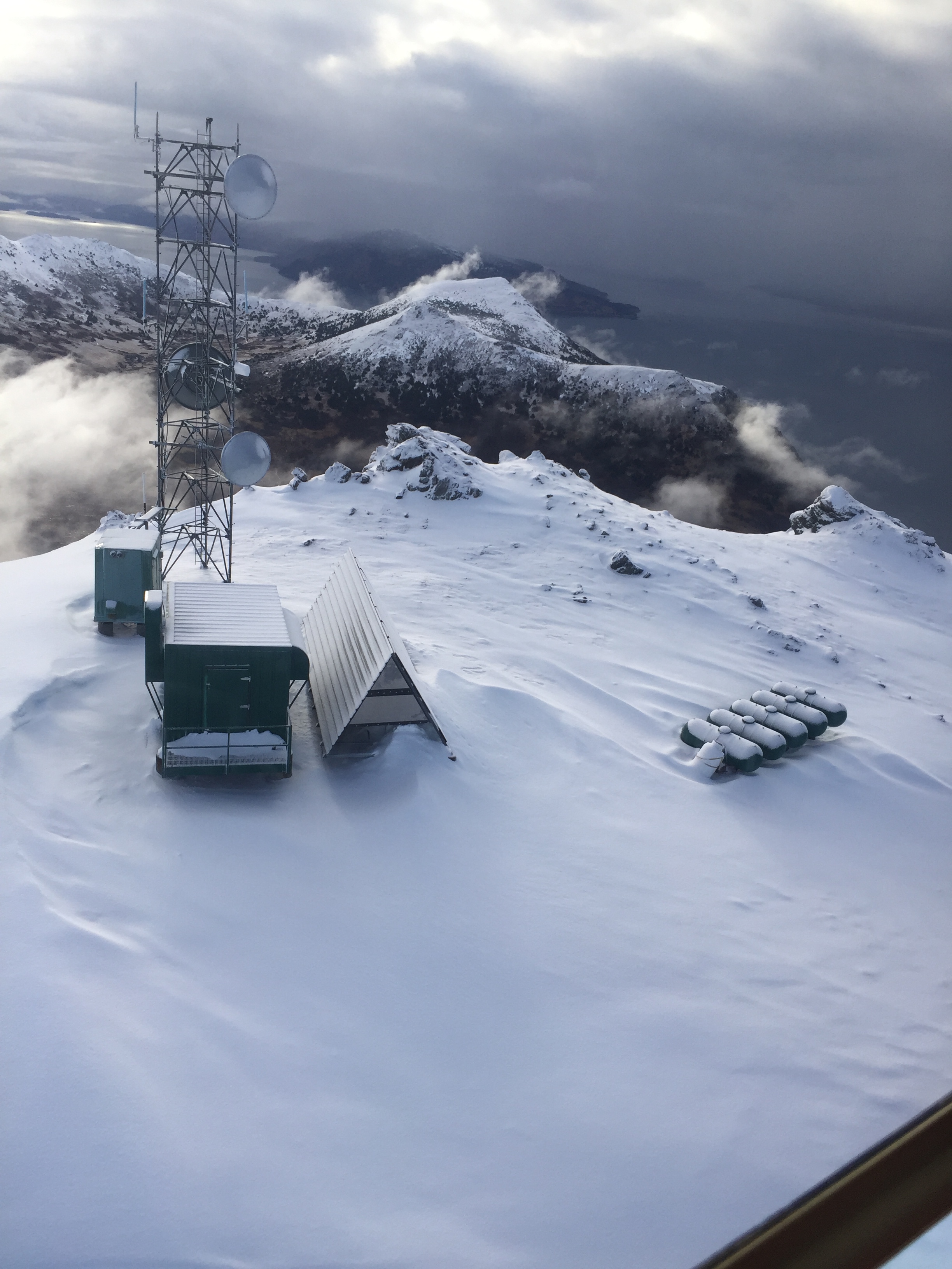 Aerial photo of snow-covered mountain with remote work site featuring a tower, 5 LP tanks, and two small buildings