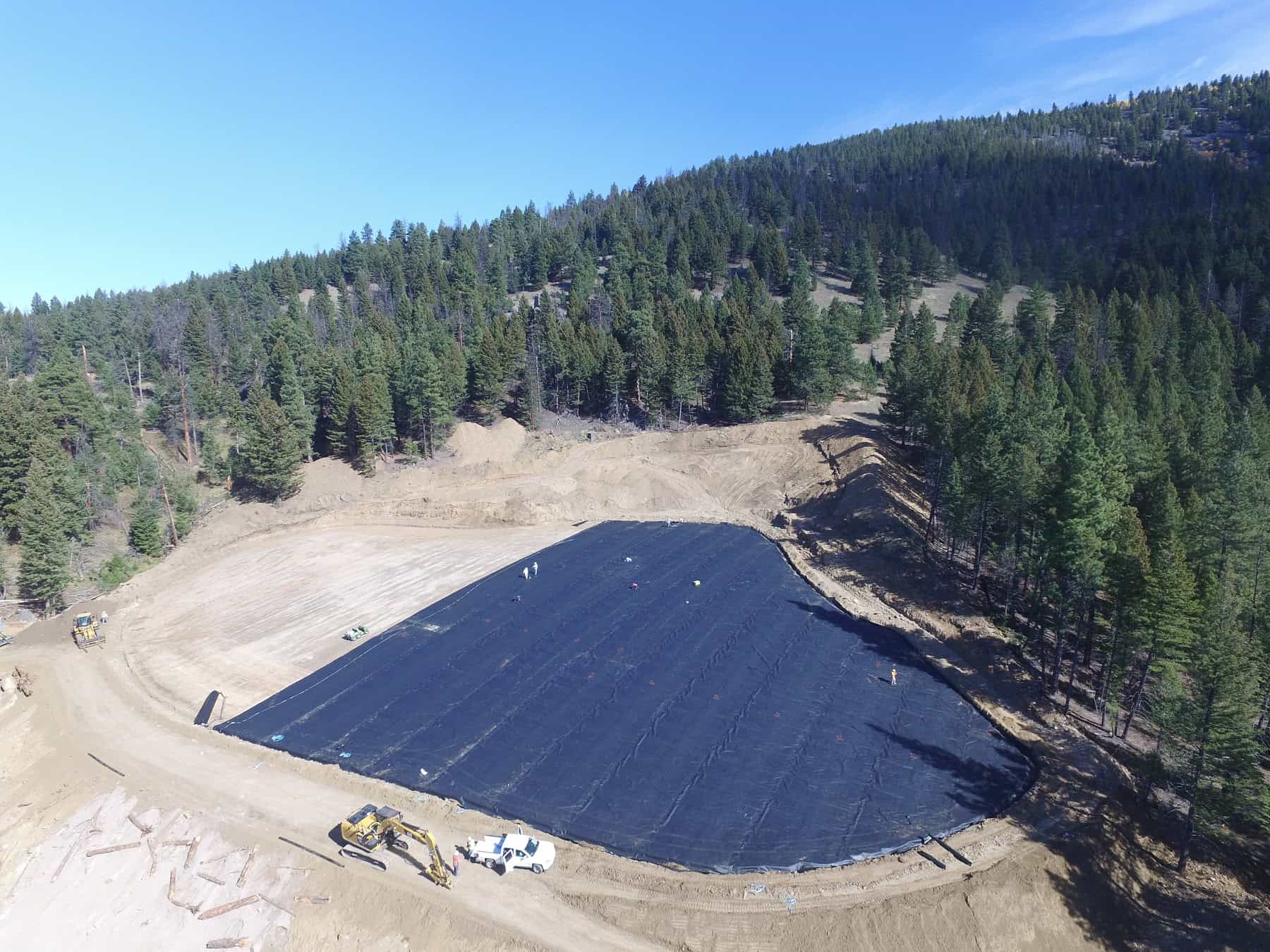 Aerial of a reclamation site in Montana with a lot of evergreen trees in the background and a large dirt area in the foreground partially covered in black covering